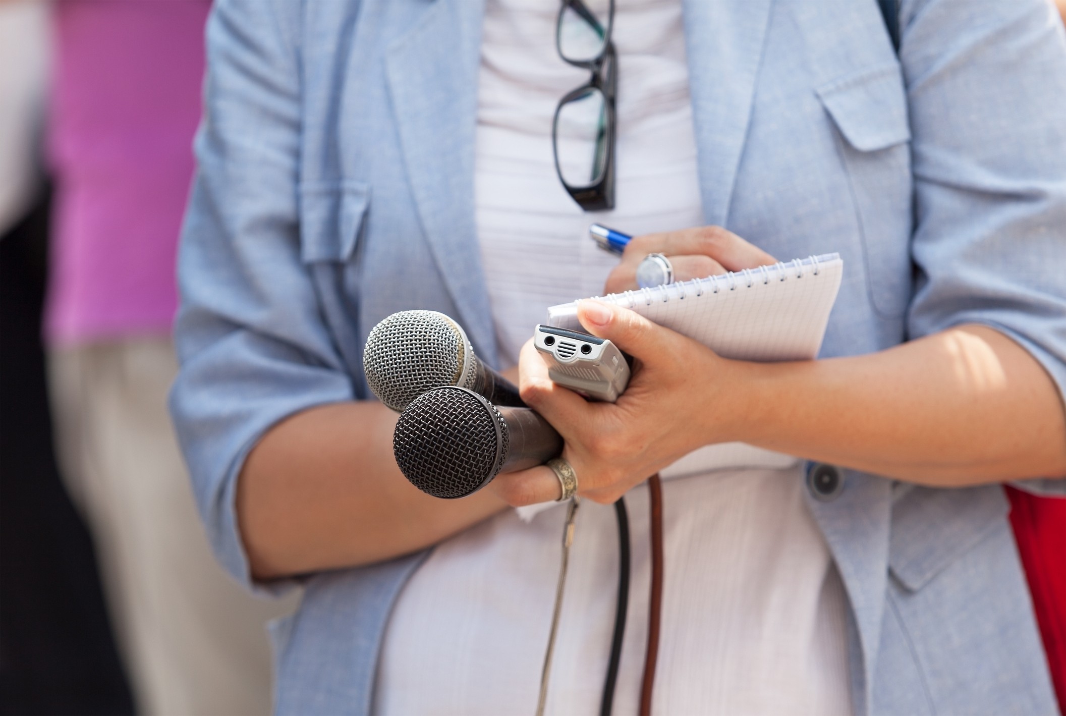 Close up of a female journalist writing on a notepad while holding microphones and a recorder at a news conference.